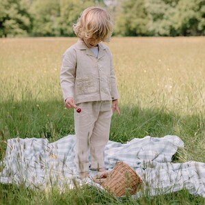 May include: A child wearing a beige linen jacket and pants set, standing in a grassy field. The jacket has embroidered details on the pockets. The child is holding a red cherry. A picnic basket and a checkered blanket are in the foreground.