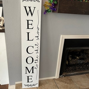 May include: White wooden sign with black lettering that reads "WELCOME to the shit show". The sign has a leafy design at the top and bottom.