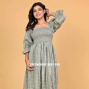 Può includere: Un lungo abito a quadretti vichy verde salvia e bianco, con scollo quadrato e maniche a tre quarti. L'abito ha un corpetto aderente e una gonna fluida. Il testo "VINTAGEHOME1959" è visibile sull'abito. La modella indossa sandali neri.