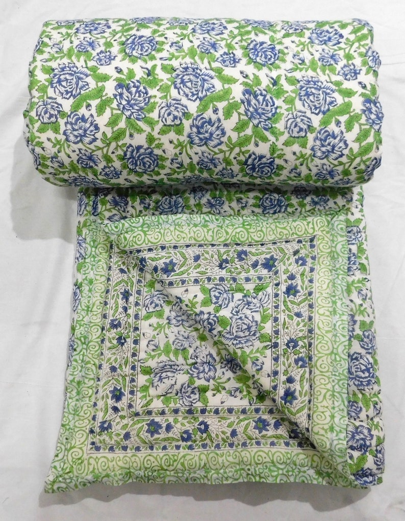May include: A white cotton quilt with a floral pattern in shades of blue and green. The quilt is folded in half and has a border of green and white.