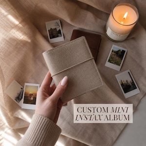 May include: A beige textured mini photo album with an elastic band closure. The album cover is plain and rectangular. The text "CUSTOM MINI INSTAX ALBUM" is printed in a dark serif font on a light rectangular overlay in the bottom right corner.