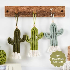 May include: Three macrame cactus wall hangings in different shades of green. The cactus are hanging from a wooden rack with hooks. The text "PDF MACRAME PATTERN" is visible in the bottom right corner of the image.