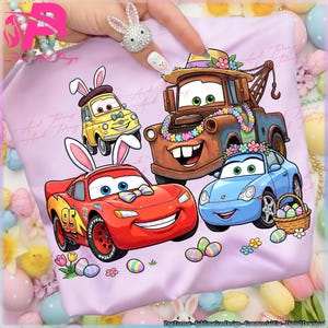 Könnte beinhalten: Ein helllila T-Shirt mit Cartoon-Autos, die mit Osterhasenohren und Dekorationen verziert sind. Zu den Autos gehören ein rotes Rennauto, ein blaues Sportauto, ein brauner Abschleppwagen und ein gelbes Auto. Ostereier und Blumen sind ebenfalls vorhanden.