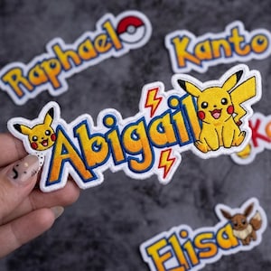 May include: Embroidered patches featuring the name "Abigail" in yellow and blue, with a Pikachu character and lightning bolt accents. Other patches include the names "Raphael", "Kanto", and "Elisa", with a Pokemon theme.
