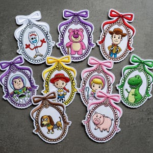 May include: A collection of embroidered patches featuring characters from the animated film series. Each patch is framed in an oval with a decorative bow at the top. Characters include a spork, a pink bear, a cowboy, and a green dinosaur.