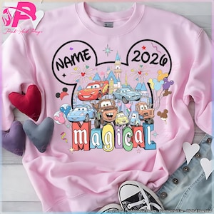 Könnte beinhalten: Ein hellrosa Sweatshirt mit einem Disney-Themen-Design. Das Design enthält das Wort "NAME" in einer Mickey-Mouse-Ohrenform, das Jahr "2026" und verschiedene Cars-Charaktere. Das Wort "magical" wird ebenfalls in bunten Blockbuchstaben angezeigt.