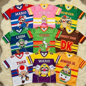 May include: Nine colorful, short-sleeved jerseys featuring characters from the Mario video game series. Each jersey has a character's name and image, including Mario, Peach, Bowser, Luigi, Yoshi, Donkey Kong, Toad, Wario, and Bowser Jr.