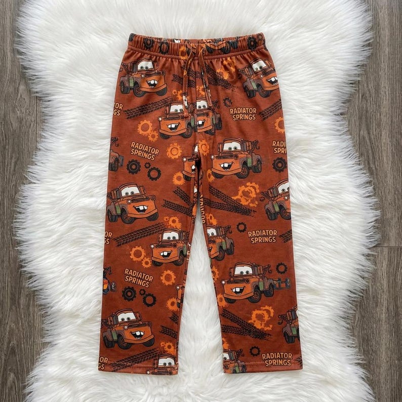 May include: Brown lounge pants featuring an all-over print of the character Mater from the movie Cars, along with the text Radiator Springs, gear icons, and tire tracks. The pants have an elastic waistband with a drawstring cord.