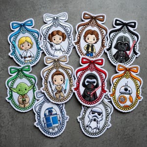 K&ouml;nnte beinhalten: Eine Sammlung von gestickten Aufn&auml;hern mit Star Wars-Charakteren in ovalen Rahmen mit dekorativen Schleifen. Zu den Charakteren geh&ouml;ren Luke Skywalker, Prinzessin Leia, Han Solo, Darth Vader, Yoda, Rey, Kylo Ren, BB-8, R2-D2 und ein Sturmtruppler.