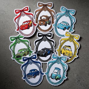 May include: A collection of embroidered patches featuring cartoon cars in oval frames with bows. Each patch has a different car design, with frames in various colors like red, brown, blue, green, black, and yellow. The patches are outlined with a white, beaded border.