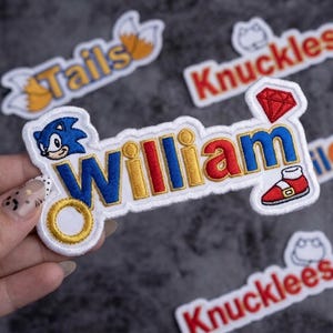 May include: Embroidered patch featuring the name "William" in blue, yellow, and red, with a cartoon Sonic the Hedgehog character and a red shoe. Other patches with the names "Tails" and "Knuckles" are also visible.