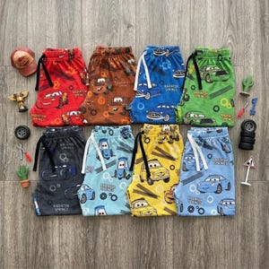 May include: Eight pairs of shorts featuring characters from the movie Cars, including Lightning McQueen and Mater, with Radiator Springs text. The shorts are red, brown, blue, green, dark gray, light blue, yellow, and light blue, each with a drawstring.