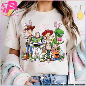 May include: Cream-colored t-shirt featuring a colorful graphic of Toy Story characters dressed for Easter, including Woody, Buzz Lightyear, Jessie, Rex, and Mr. Potato Head, all wearing bunny ears. Easter eggs and baskets are also included in the design.