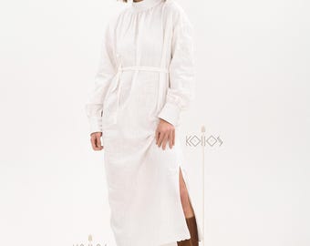 Puede incluir: Un vestido blanco de manga larga con cuello alto y cinturón de lazo. El vestido tiene una abertura lateral y un dobladillo festoneado. La modelo lleva botas marrones de caña alta.