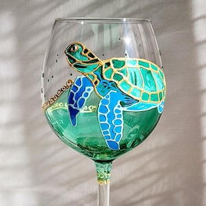 Könnte beinhalten: Ein grünes Weinglas mit handbemaltem Design einer Meeresschildkröte. Die Schildkröte ist grün und blau mit goldenen Akzenten. Das Glas steht auf einer Holzoberfläche.