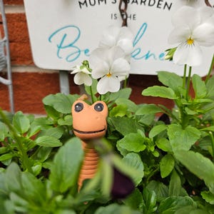 May include: A brown ceramic garden gnome shaped like a worm with a smiling face peeking out from a pot of white pansies. The sign behind the gnome reads "Mum's Garden is the Best".