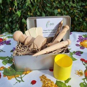 May include: A silver tin filled with wooden tools for making paper pots. The tools include a paper pot maker, a wooden dibble, and a wooden trowel. The tin also includes a yellow paper pot. The tin has a label that says "O-pots Paper Pot Maker & Wooden Tools Gift Set".
