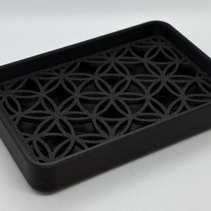 K&ouml;nnte beinhalten: Eine schwarze Seifenschale mit einem geometrischen Muster, das aus dem Boden herausgeschnitten ist. Die Schale ist aus hartem Kunststoff.