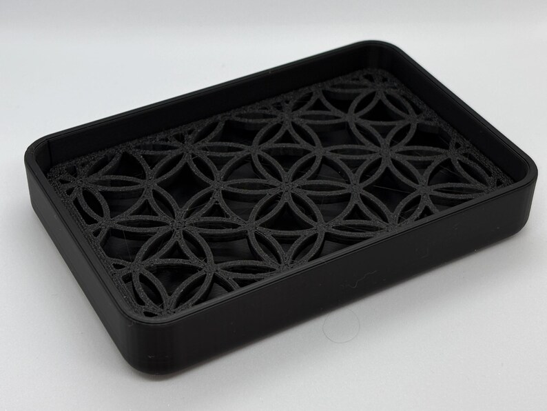 K&ouml;nnte beinhalten: Eine schwarze Seifenschale mit einem geometrischen Muster, das aus dem Boden herausgeschnitten ist. Die Schale ist aus hartem Kunststoff.