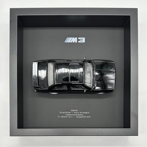 May include: A black BMW M3 model car in a shadow box frame. The car is displayed on a grey background with the text "BMW M3" and specifications: 215 pk/125 kW | 2.02 cc 16V straight 4 | 5-speed manual gearbox | 0-100 km/h 6.7 s. | top speed 241 km/h.