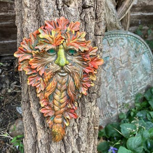 May include: A colorful Green Man face sculpture with green eyes, a green nose, and a long, flowing beard. The sculpture is attached to a tree trunk. The leaves around the face are orange, yellow, and green.