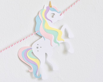 Peut inclure: Une découpe de licorne en papier blanc avec une crinière et une queue arc-en-ciel. La licorne est suspendue à une ficelle rouge et blanche.