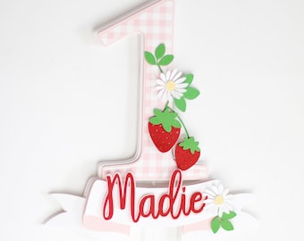 May include: A pink and white gingham number one cake topper with red strawberries, white daisies, and green leaves. The name "Madie" is written in red script on a white banner. A festive decoration for a first birthday.