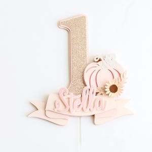 May include: A light pink and gold glitter number one cake topper with a pumpkin and sunflower design. The name "Stella" is written on a banner. The cake topper is designed for a first birthday celebration.