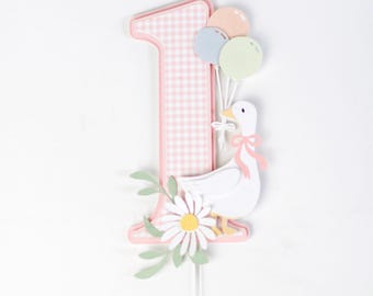 May include: A pink and white gingham number one cake topper with a white goose holding balloons. The goose is wearing a pink bow and the balloons are pastel pink, blue, and green. The topper is decorated with green leaves and a white daisy.