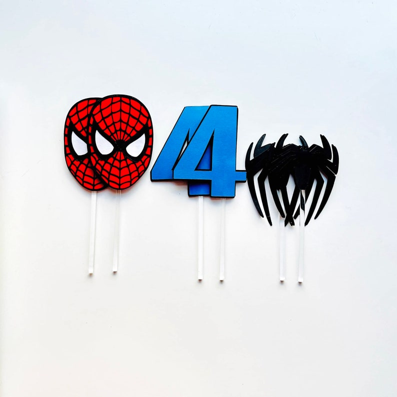 May include: Three cake toppers featuring a red and black Spiderman mask, a blue number four, and a black spider. The toppers are on white sticks.