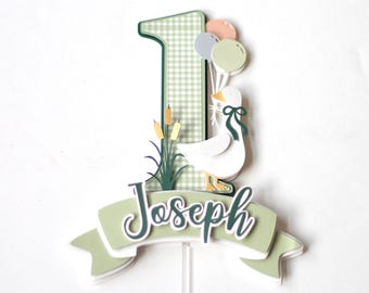 May include: Green and white gingham number one cake topper with a white duck holding balloons and a green banner with the name Joseph.