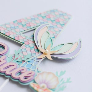 May include: A layered mermaid-themed cake topper in pastel shades. The design features a mermaid tail, a seashell, and the name "Grace". The number "4" is also present, suggesting a birthday theme. The topper is made of paper.