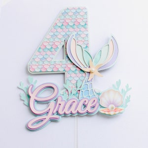 May include: A layered paper cake topper with a mermaid theme, featuring the number 4 and the name "Grace". The number has a scale pattern in pastel colours. The design includes a mermaid tail, a seashell, and a starfish.