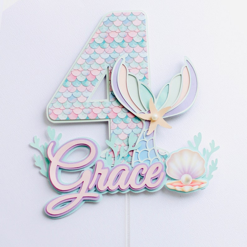 May include: A layered paper cake topper with a mermaid theme, featuring the number 4 and the name "Grace". The number has a scale pattern in pastel colours. The design includes a mermaid tail, a seashell, and a starfish.