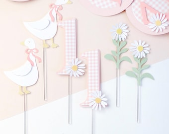 May include: Party decorations with a pink and white gingham design. The set includes the number "1" with daisy accents, paper geese with pink bows, and daisy flower picks. The colour scheme is soft pink, white, and yellow, perfect for a first birthday celebration.