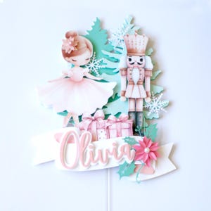 May include: A pink and green cake topper featuring a ballerina and a nutcracker. The topper includes a white banner with the name "Olivia" in pink lettering. The topper is perfect for a Christmas or winter-themed birthday party.