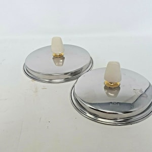 Puede incluir: Dos tapas de olla plateadas con pomos blancos y detalles dorados. Las tapas son redondas y tienen un borde elevado.