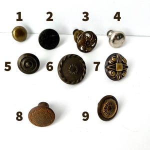 May include: A collection of nine distinct metal cabinet knobs arranged in a grid, numbered one through nine. The knobs feature various finishes including brass, black, silver, and antique bronze, with diverse textures, floral patterns, and circular designs.