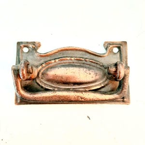 May include: A rectangular metal drawer pull with a weathered copper finish. The hardware features a central oval plate with a decorative border and a hinged, U-shaped handle attached to two circular mounts. Two screw holes are visible at the top corners.