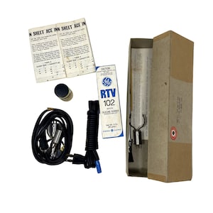 Puede incluir: Un kit de sellador de caucho de silicona blanco RTV 102 de General Electric vintage en una caja de cartón marrón. El kit incluye un tubo de sellador, una pequeña botella de líquido, un par de alicates, una pequeña herramienta de metal y un cable negro con un enchufe azul.