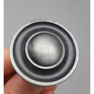 May include: A circular metal cabinet knob with a dark, brushed gunmetal gray finish. The knob features a raised, smooth central dome surrounded by a recessed circular channel and a flat, wide outer rim, creating a layered, concentric design.