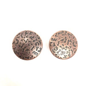 Peut inclure: Une paire de boucles d'oreilles rondes en cuivre avec une surface texturée et à motifs. Les boucles d'oreilles sont de couleur cuivre foncé avec un motif noir.