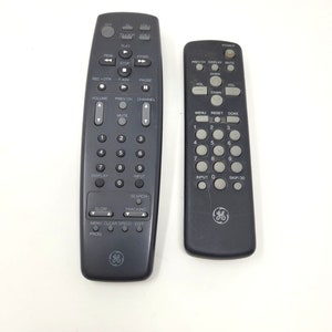 Puede incluir: Dos mandos a distancia negros con botones para funciones de TV y VCR. El mando de la izquierda tiene botones para reproducir, detener, rebobinar, avanzar rápido, pausar, grabar, subir y bajar el canal, subir y bajar el volumen, mostrar, entrada, buscar, lento, seguimiento, menú, borrar, velocidad, editar y prog. El mando de la derecha tiene botones para encender, canal anterior, mostrar, silenciar, canal, volumen, menú, restablecer, hecho, 1-9, entrada, saltar 30 y 0.