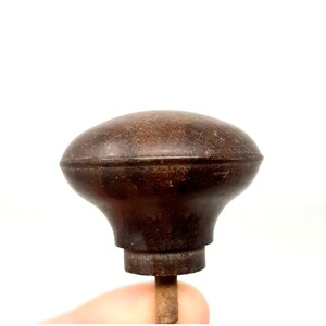 May include: A dark brown, rounded door knob with a smooth, slightly weathered surface and a central horizontal ridge. The knob features a flared, mushroom-like shape that tapers down to a cylindrical base, with a metal spindle extending from the bottom.