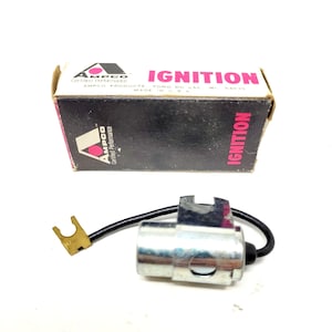 May include: A black box with white text that reads "AMPCO Certified Performance IGNITION AMPCO PRODUCTS FOND DU LAC WI 54935 MADE IN USA" and a red triangle with a white circle inside. The box is sitting on a white surface. A silver ignition condenser with a black wire and a gold connector is also on the white surface.