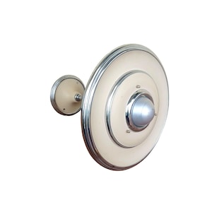 May include: A wall-mounted light fixture featuring a large, circular, cream-colored disc with concentric chrome rings and a central silver dome. A smaller, matching circular cream-colored base connects the fixture to the wall surface.