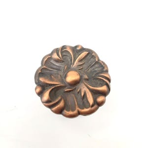 May include: A round cabinet knob with a copper-toned finish and a dark, recessed patina. The surface features a raised, symmetrical floral-inspired relief pattern with a central circular dome and radiating petal-like segments around the edge.