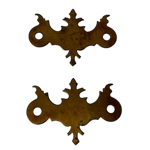 Op de afbeelding: Twee bijpassende ladeplaten van messing met een donkere, verouderde patina. Elke plaat heeft een centraal verticaal punt, symmetrische zijrollen en twee ronde gaten aan de buitenste uiteinden voor bevestigingsmateriaal. Het metaal vertoont slijtage.