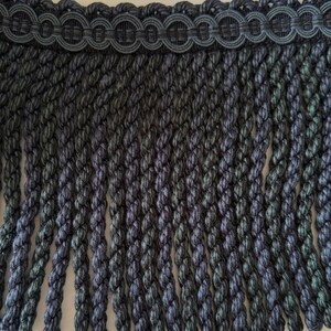 May include: Close-up of a decorative fringe trim in dark grey and green hues. The trim has a woven top with a repeating circular design, and long, braided fringe hanging below. Ideal for home decor and craft projects.