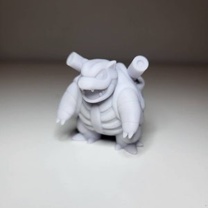 May include: A light gray figurine of a Blastoise character, a Pokémon. The figure has a turtle-like appearance with a large shell, two cannons on its back, and a smiling expression. The figure is on a white surface.